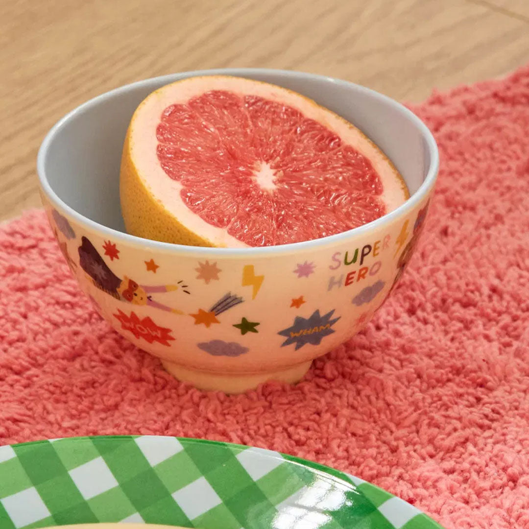 rice kinderkom met hero-thema, gevuld met een halve grapefruit, staand op een roze kleed.