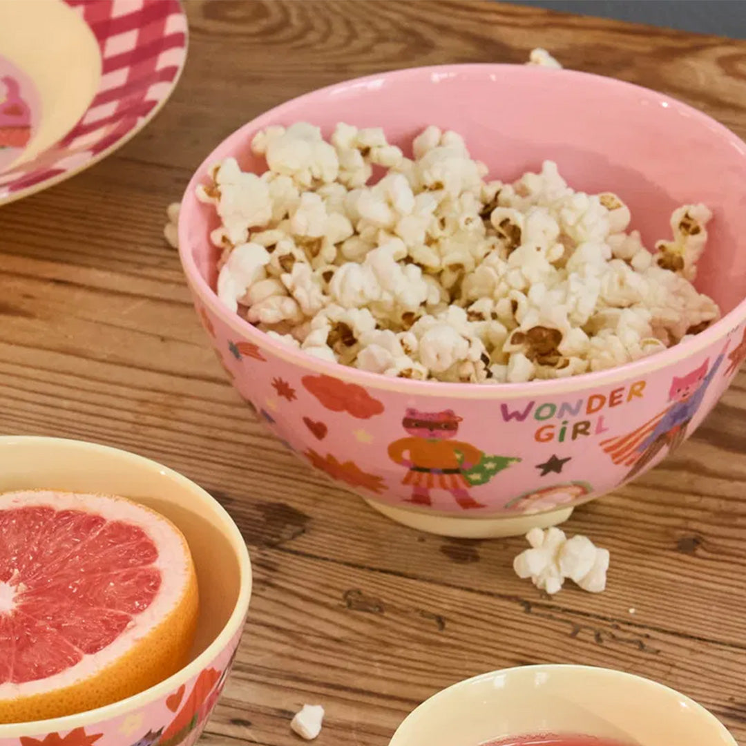 Roze rice kinderkom met superheldenprint, gevuld met popcorn op een houten ondergrond.
