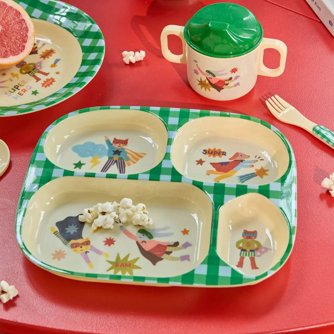 rice vakjesbord voor kinderen met superheldenfiguren, een bijpassende beker en popcorn op een rode tafel.