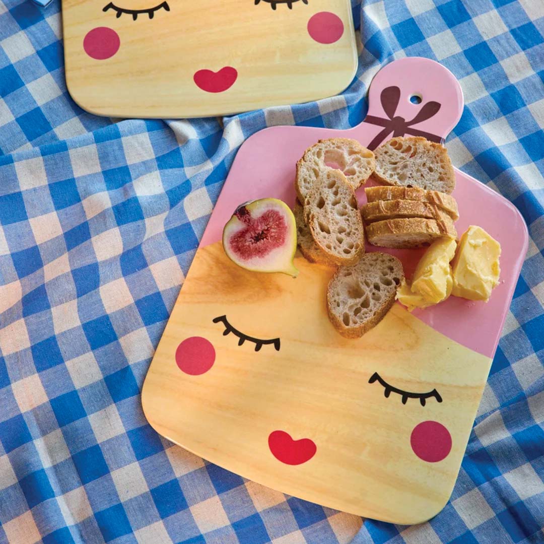 Twee snijplanken met gezichtsdesign op een blauw-wit geruite doek, waarvan één met brood, boter en een vijg.