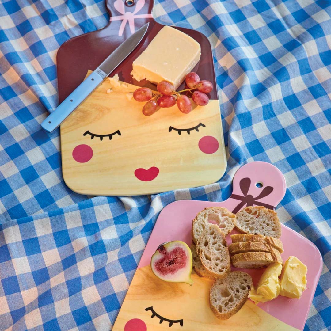 Twee Rice snijplanken met 'lady' gezichtspatroon, één bruin en één roze, met hapjes op een blauw-wit geruite doek.