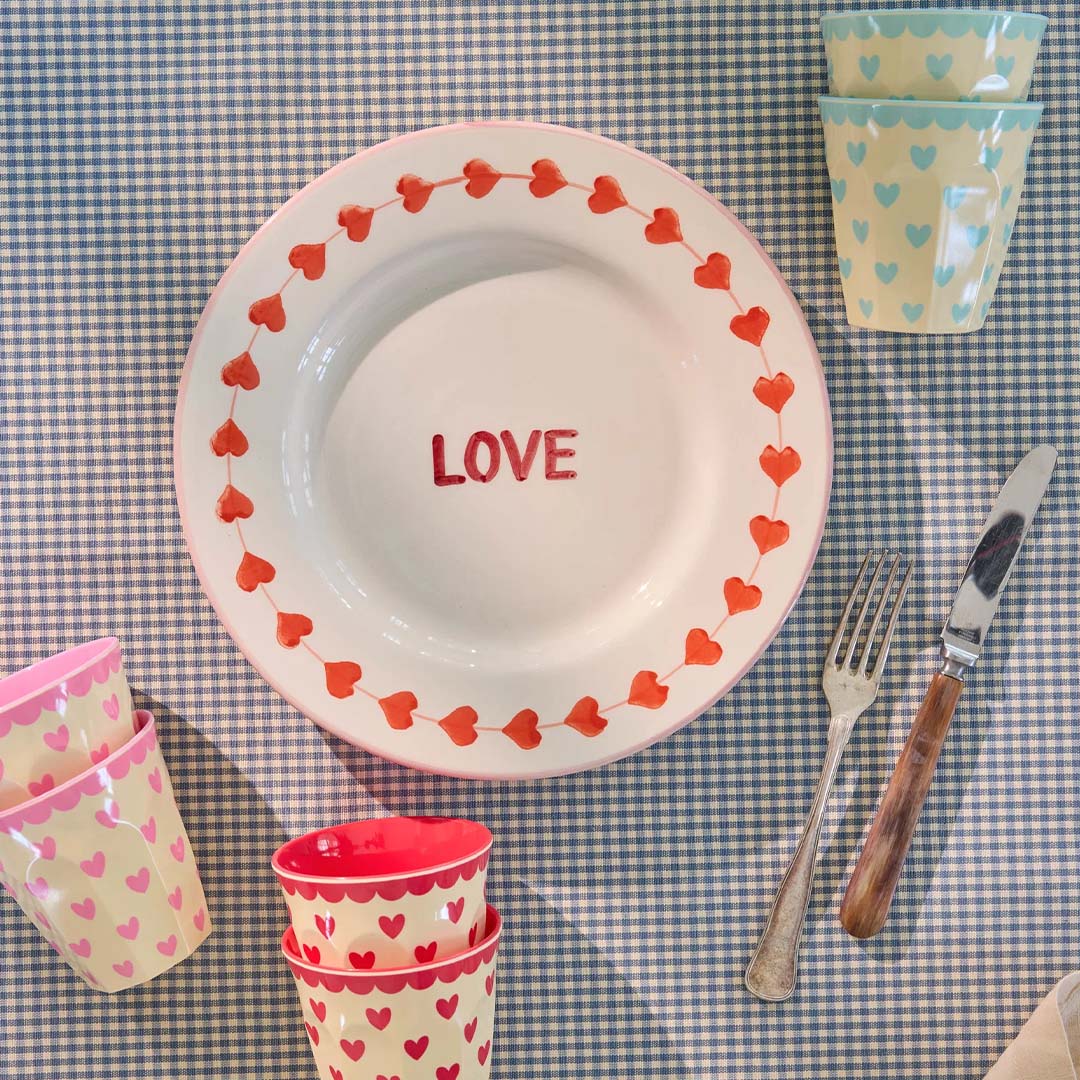 Tafelsetting met Rice kinderbekers met hartenpatroon, een bord met 'LOVE' en bestek op een blauw-wit geruite tafelkleed.