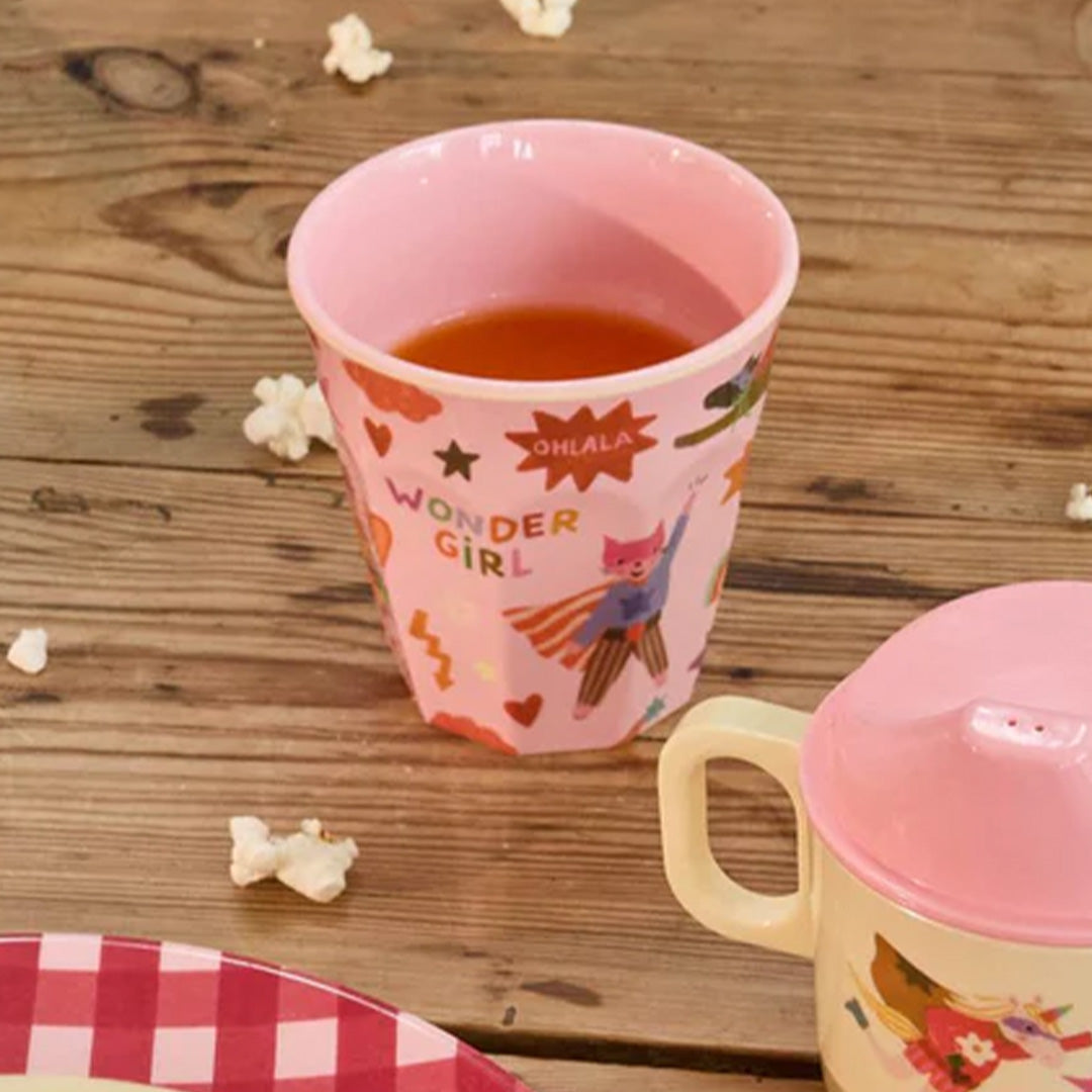 Een roze rice kinderbeker met superheldenkat, gevuld met drinken, staat op een houten tafel met popcorn.