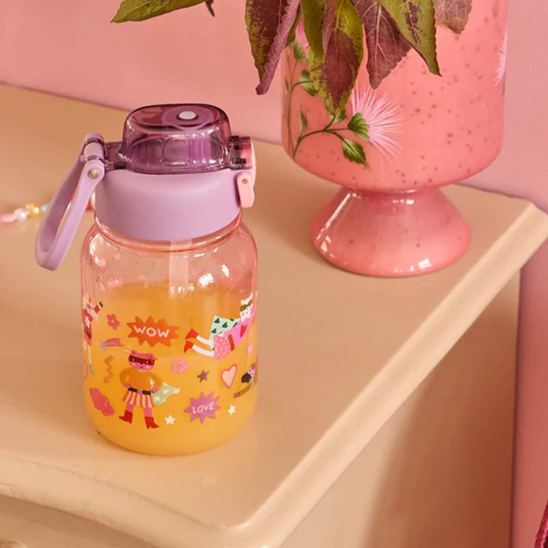 Kinderdrinkfles met superheldenprint, gevuld met oranje drank, op een tafel naast een roze vaas.