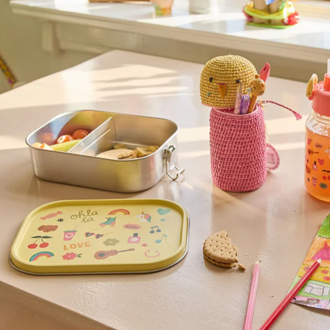 rice RVS broodtrommel met gele deksel op een tafel, gevuld met fruit en koekjes, naast een roze pennenhouder.
