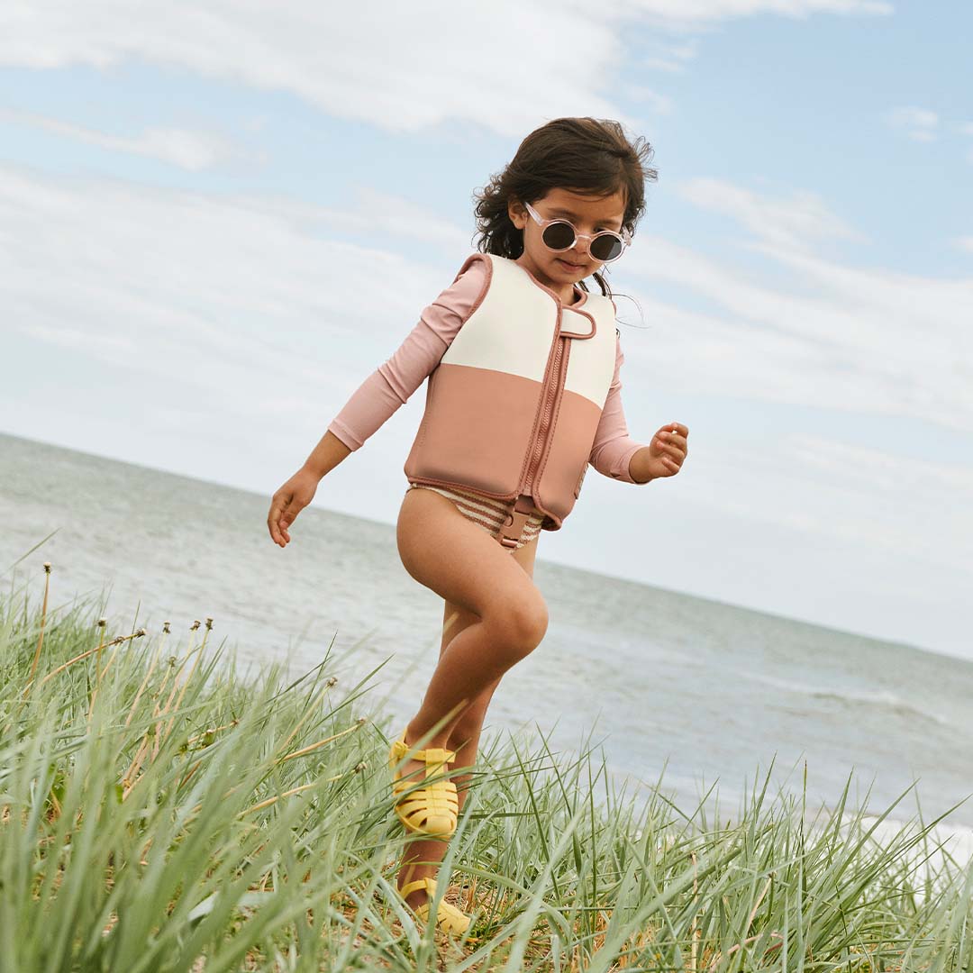 Een kind met een roze en crème zwemvest en zonnebril loopt door het gras bij de zee.