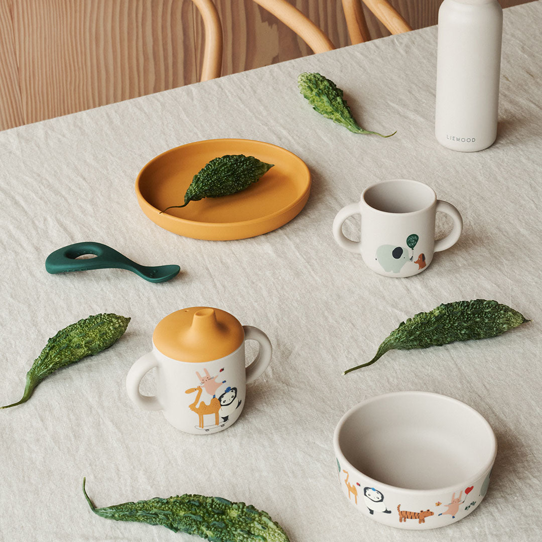 Liewood siliconen kinderservies met twee bekers, een gele plaat, een kom met dierenpatroon en een groene lepel op een tafel.