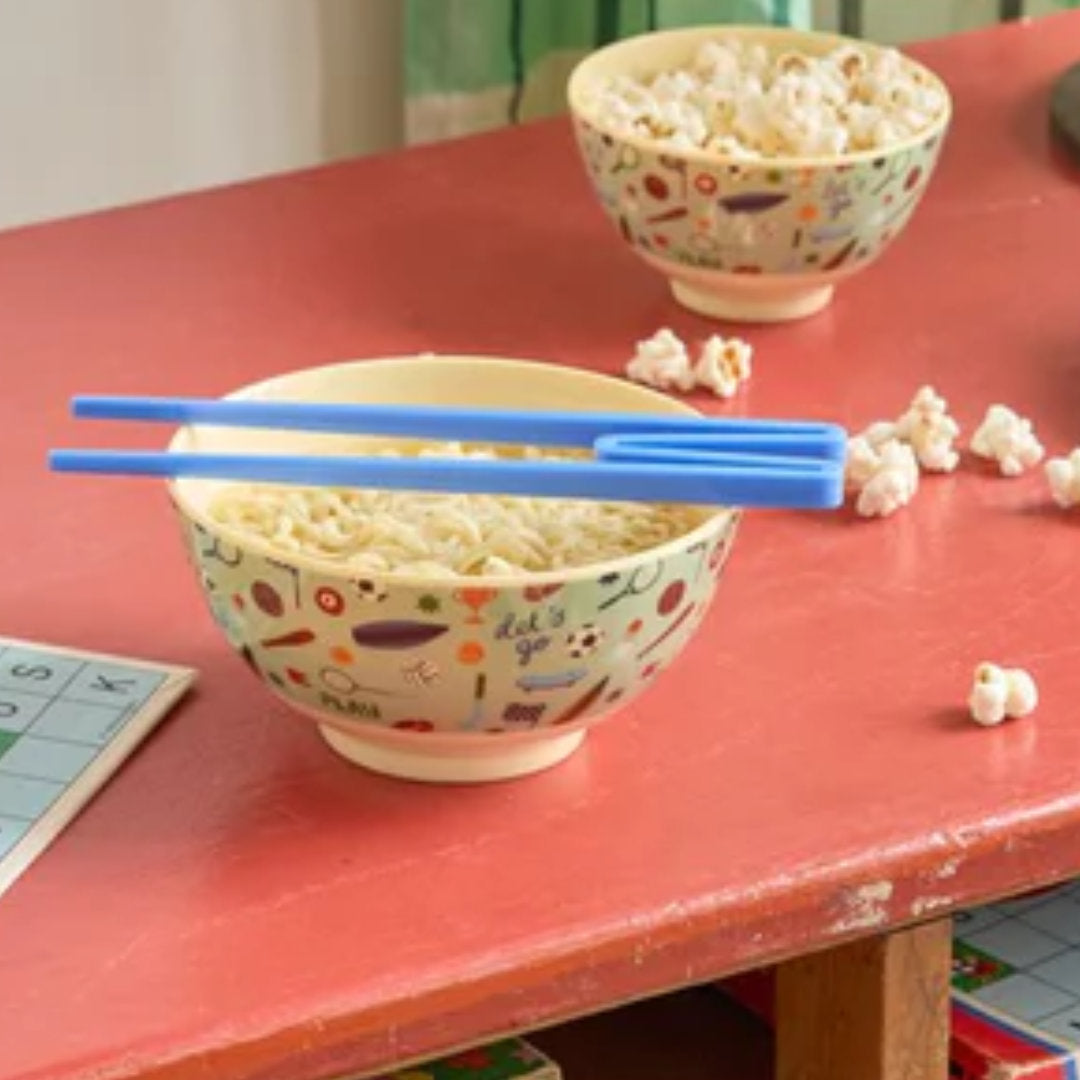 Twee rice kinderkommen met sportthema op een rode tafel, één met noodles en blauwe eetstokjes, de andere met popcorn.