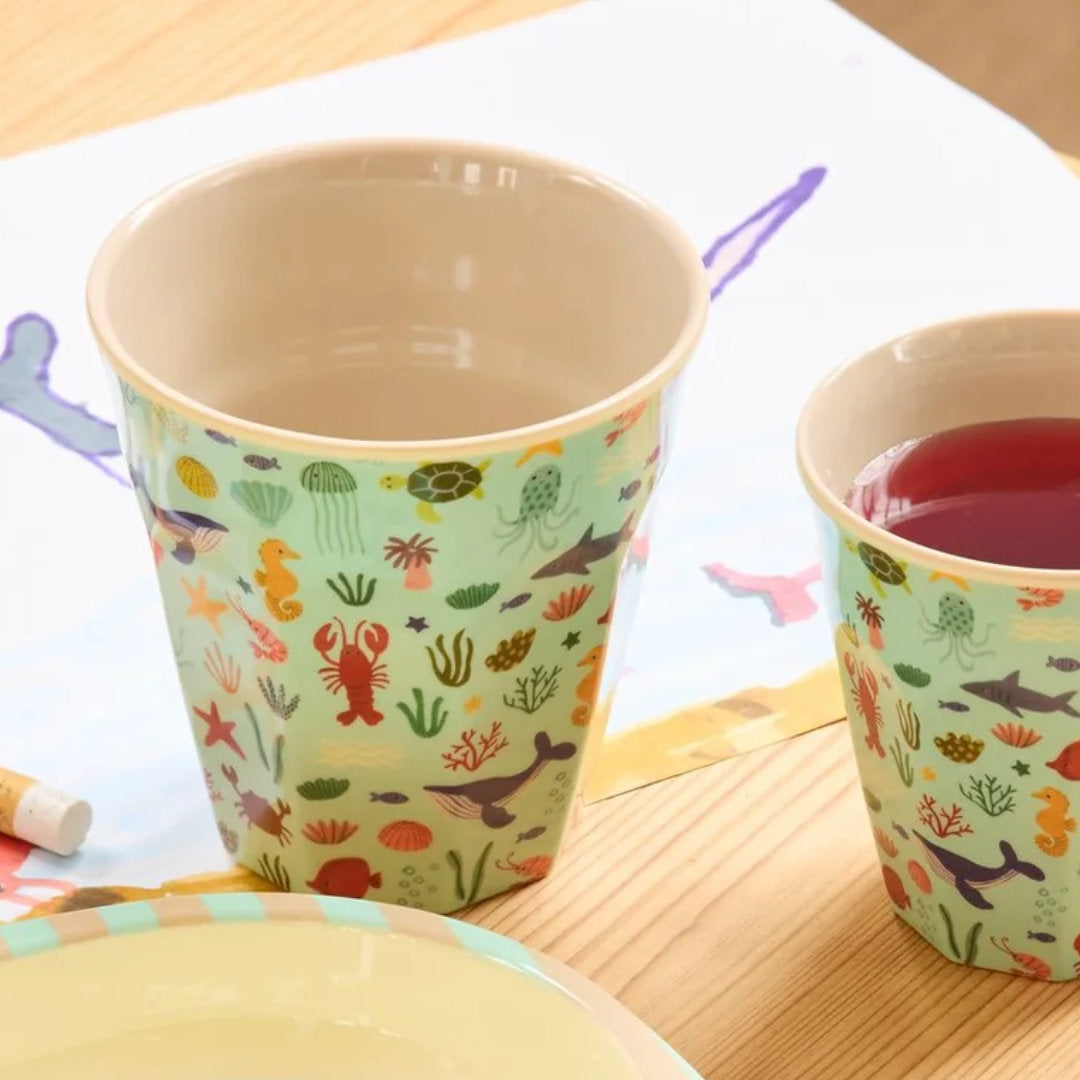 Twee Rice kinderbekers met zeedierenpatroon, waarvan één gevuld met rode drank, op een houten tafel naast een tekening.