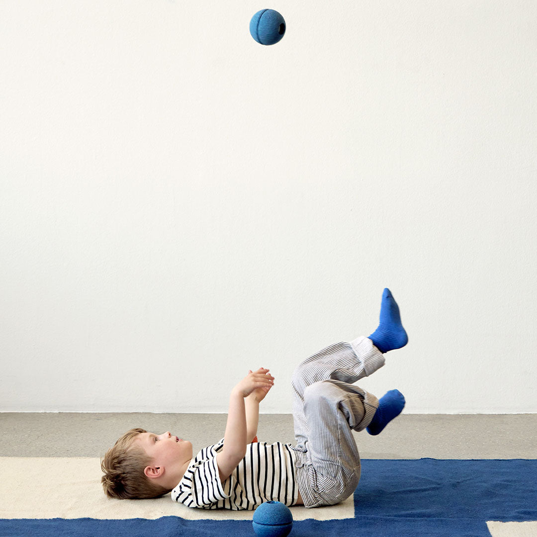Een kind ligt op de rug en speelt met een Modu diepblauwe schuimrubberen bal die in de lucht is.