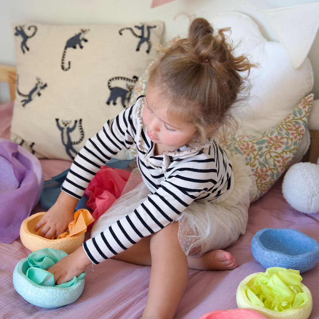 Een kind speelt met verschillende gekleurde pit and play speeldoeken in ronde bakjes op een bed.