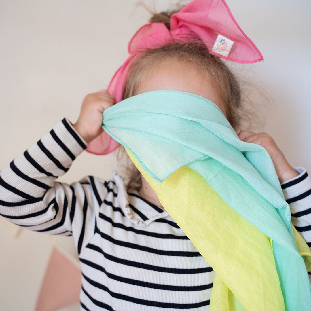 Een kind speelt kiekeboe met een pastelgroen en geel speeldoek.