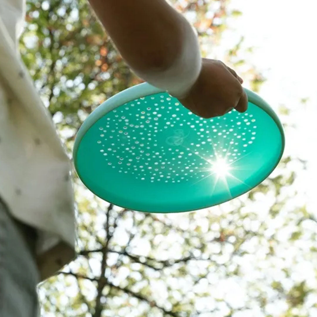 Een kind houdt de Quut blauwe frisbee en zandzeef omhoog tegen de zon, met bomen op de achtergrond.