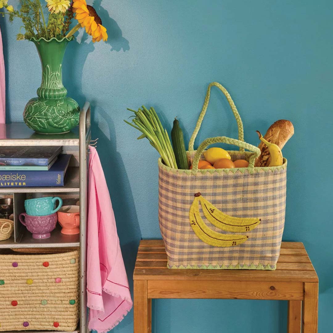 De geruite raffia kinderhandtas met bananen, gevuld met boodschappen, op een houten bankje.