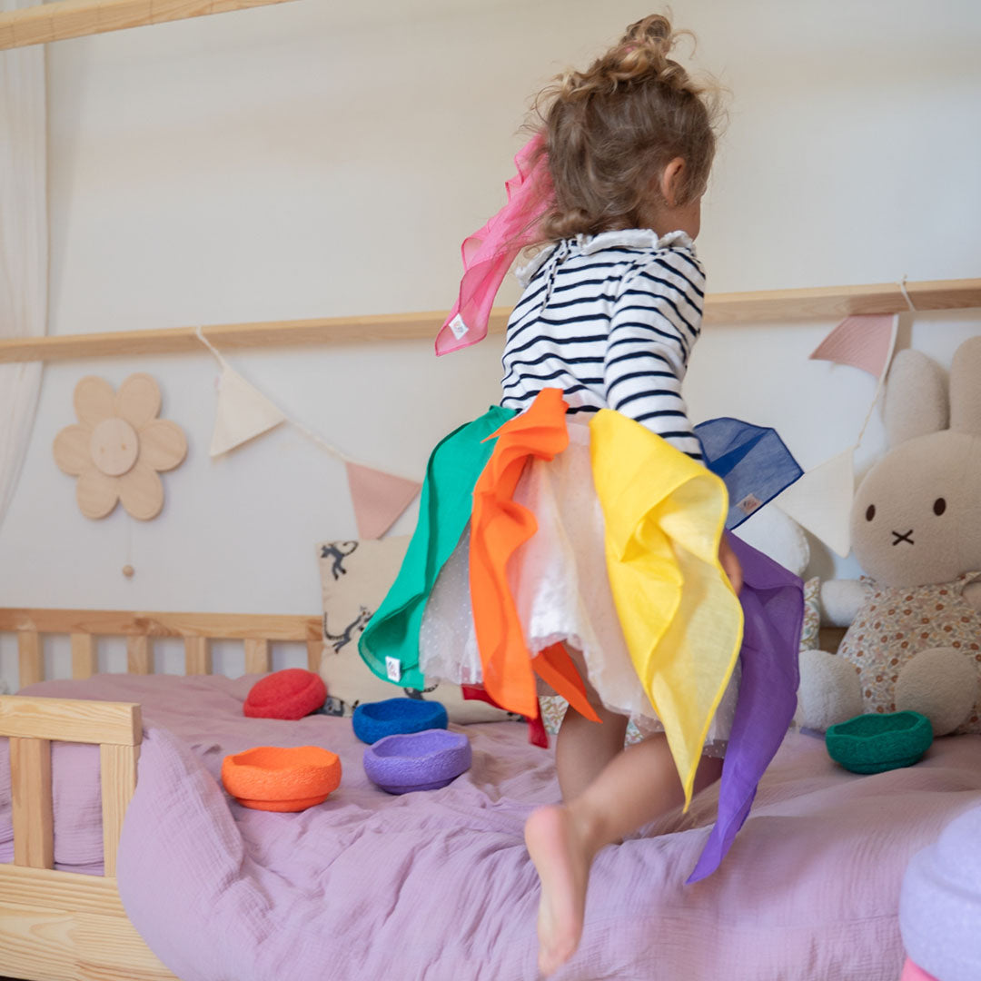 Een kind springt op bed met regenboogkleurige speeldoeken als rok.