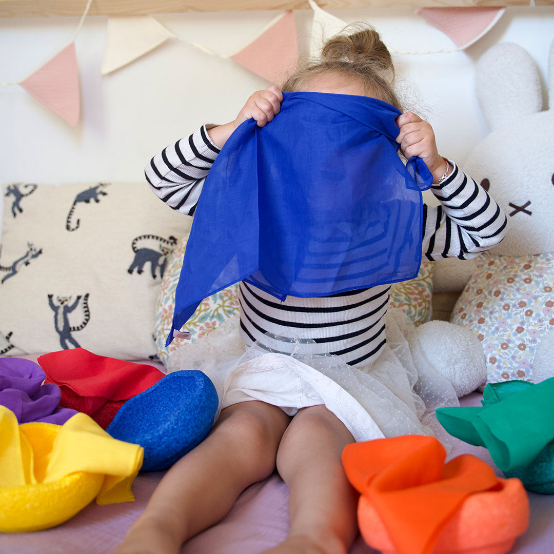 Een kind speelt kiekeboe met een blauwe pit and play speeldoek op een bed, omringd door andere kleuren.