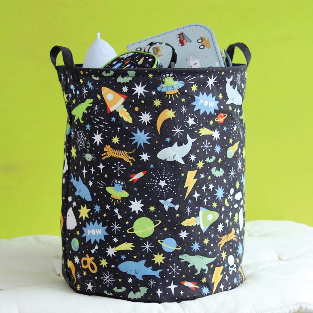 A Little Lovely Company opbergmand met galaxy print, gevuld met spullen, op een wit kussen voor een groene achtergrond.