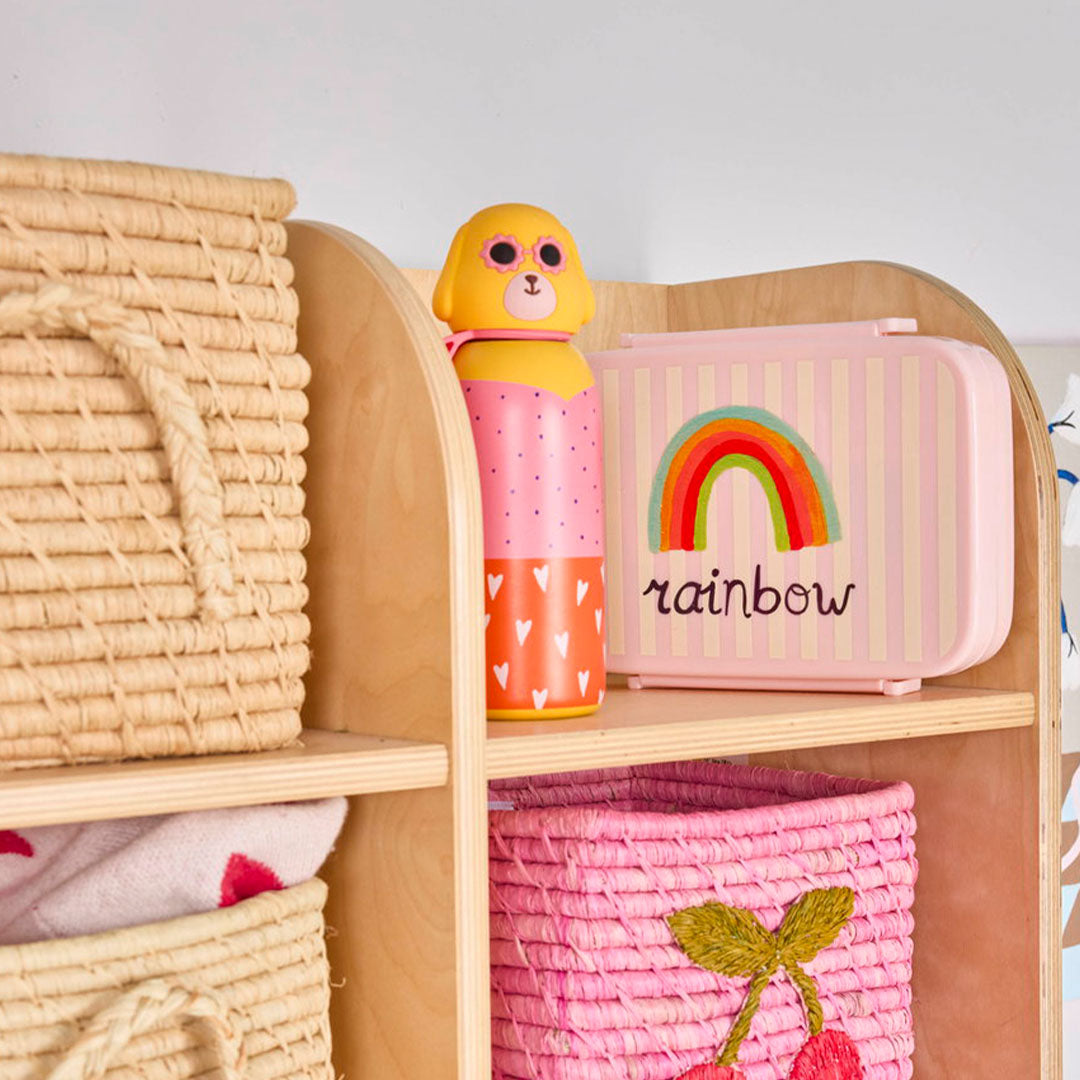 De rice rvs thermos drinkfles met hondenhoofd staat op een houten plank naast een roze lunchbox met een regenboog.