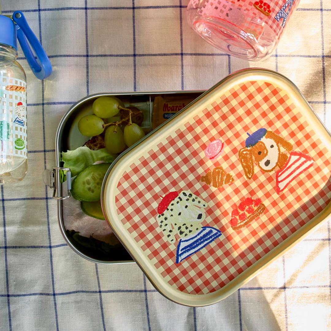 Open rice rvs kinderbroodtrommel gevuld met lunch, met deksel met Parijse hondenfiguren ernaast.