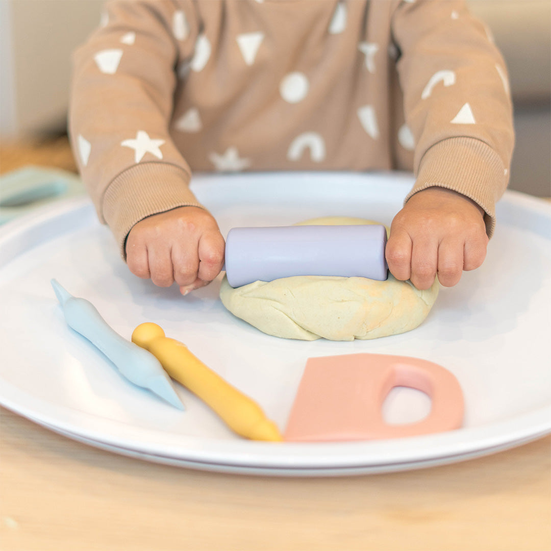 Kinderhanden rollen klei met een paarse siliconen deegroller uit de Inspire My Play set op een witte schaal.