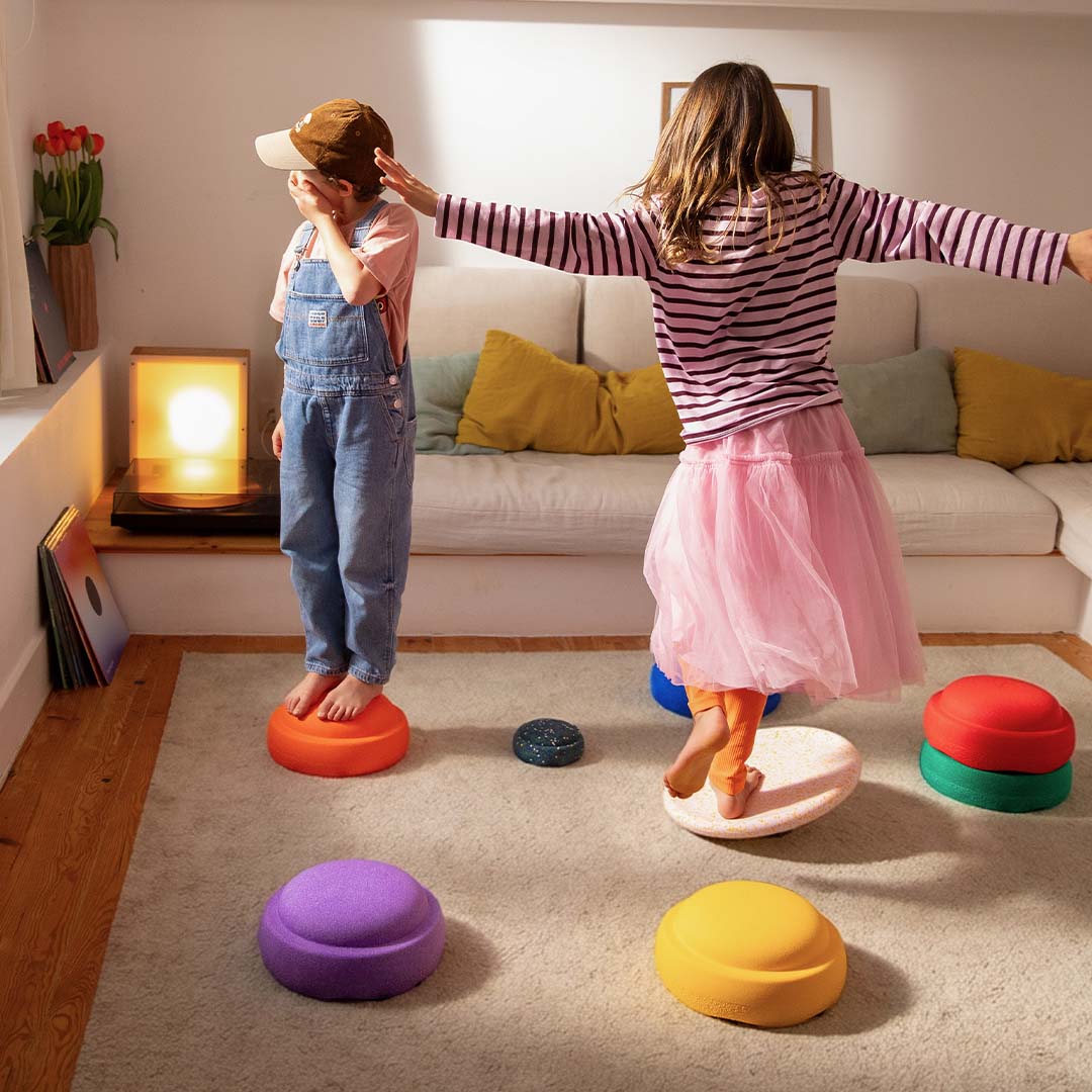 Twee kinderen spelen binnenshuis met stapelstein balansspeelgoed, staand op de gekleurde stapelstenen.