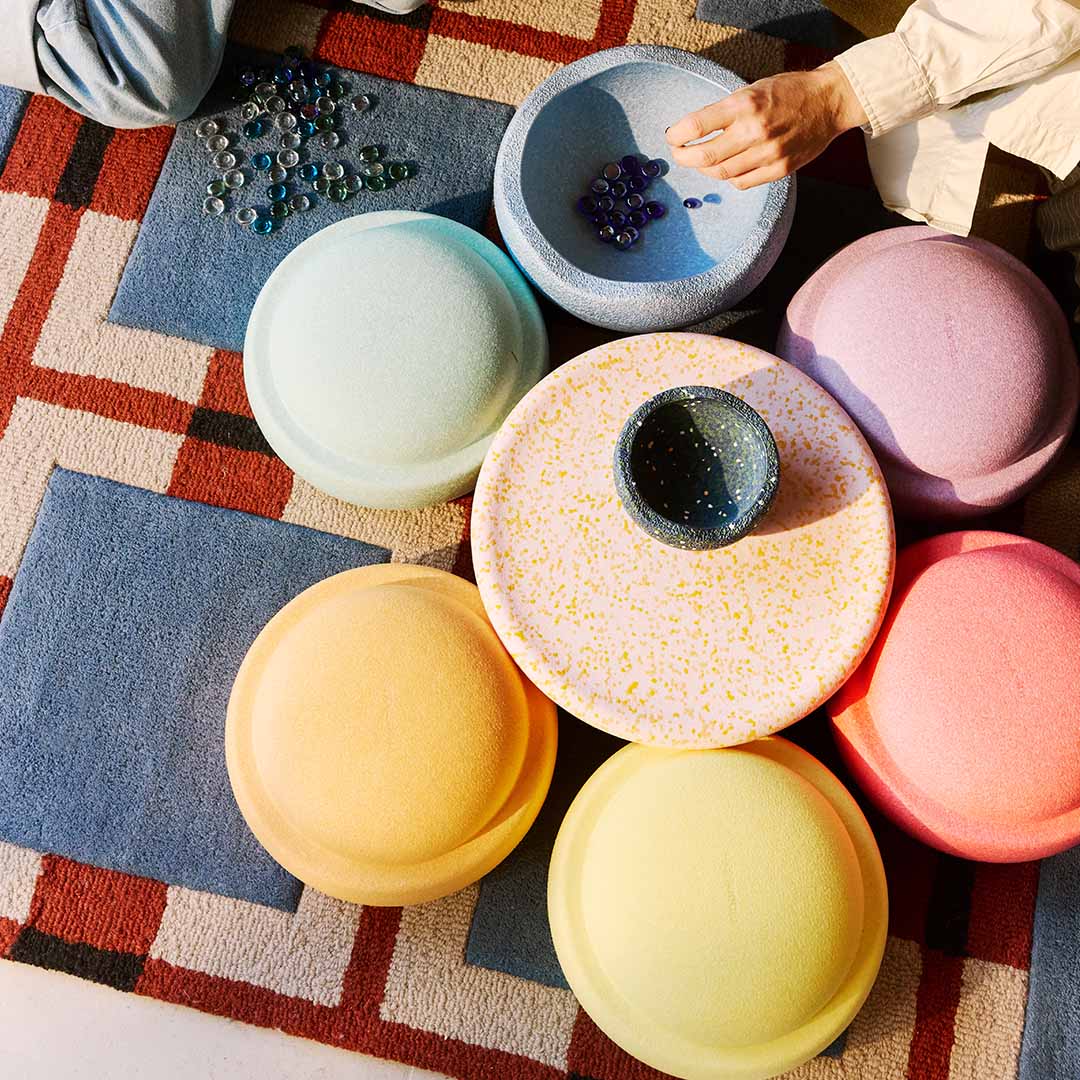 Bovenaanzicht van stapelstein speelgoed in pastelkleuren op een vloerkleed, met een hand die knikkers in een van de elementen legt.