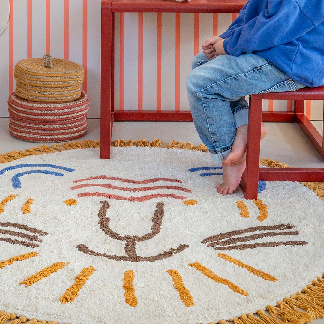 Detail van het ronde vloerkleed met leeuwenkop en de blote voeten van een kind dat ernaast zit.