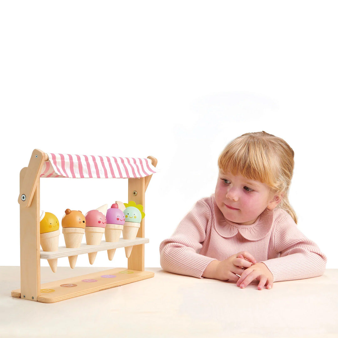Een meisje in een roze trui zit naast de Tender Leaf Toys houten ijswinkel speelset.