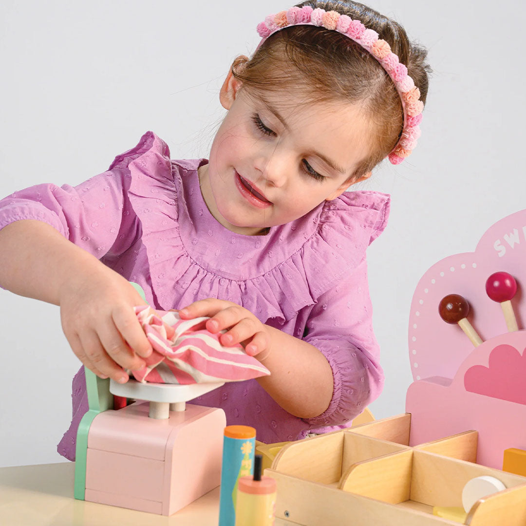 Een meisje weegt een gestreepte doek op de weegschaal van de Tender Leaf Toys houten speelset snoepwinkel.