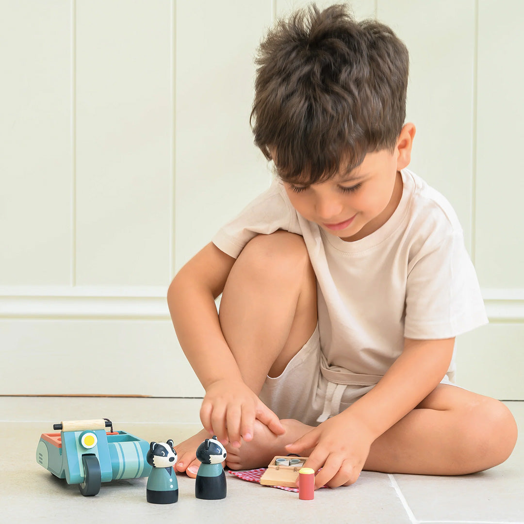 Een jongen speelt binnen op de grond met de Tender Leaf Toys houten speelset van dassen op motor en picknickspullen.