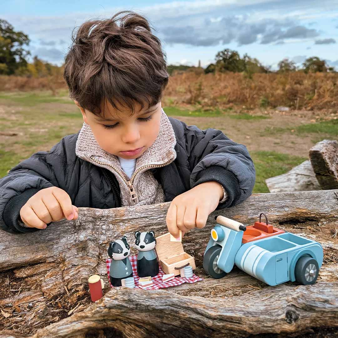 Een jongen speelt buiten met de Tender Leaf Toys houten speelset van dassen op motor en picknickspullen.