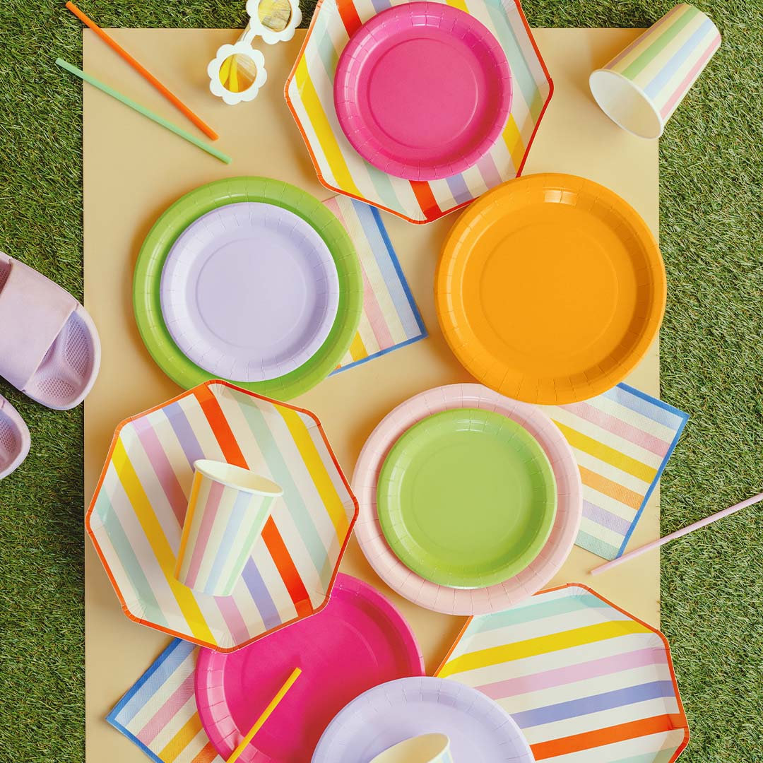Partydeco collectie van veelkleurige gestreepte en effen papieren feestborden, bekers en servetten op een beige ondergrond.