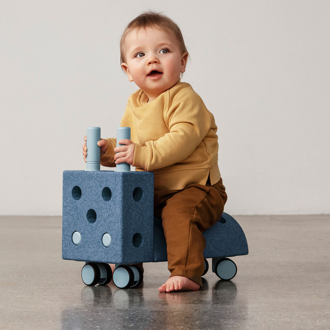 Baby zit op een Modu blauw schuimrubberen speelgoedvoertuig met wielen en handvatten.