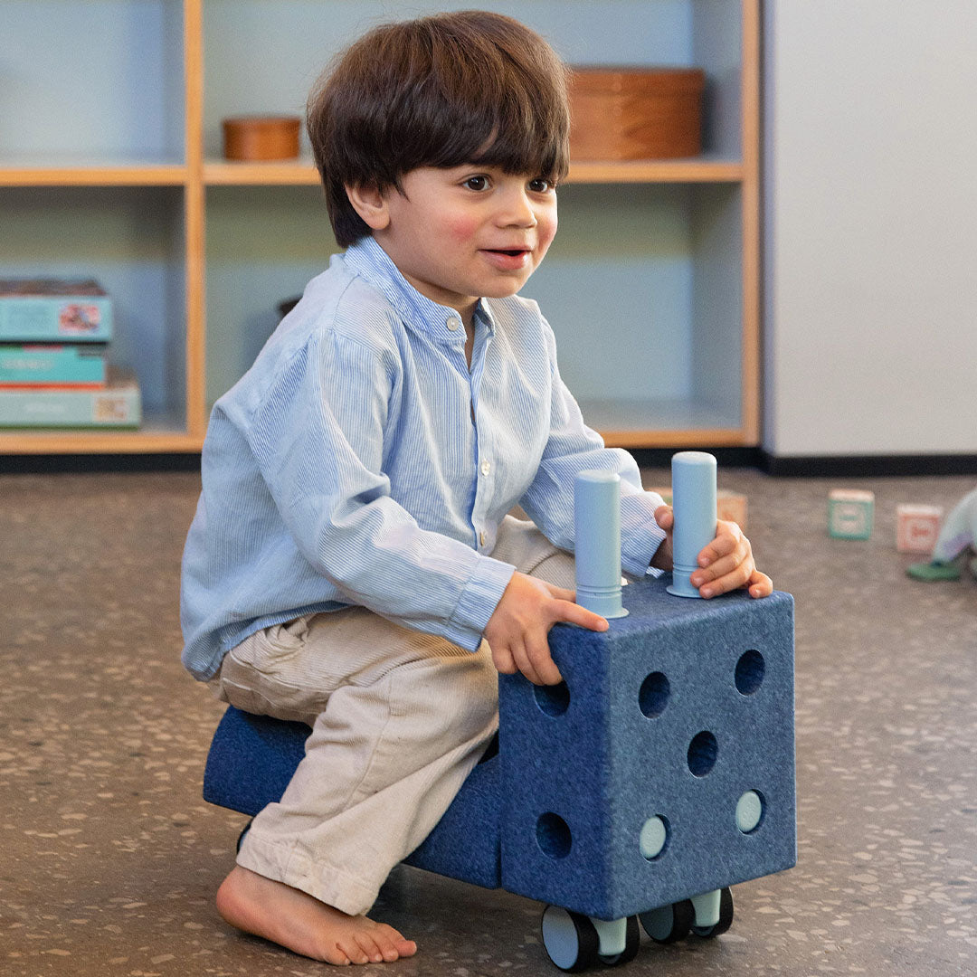 Een jongen zit op een blauw modu rijdend speelgoedvoertuig met twee lichtblauwe handvatten.