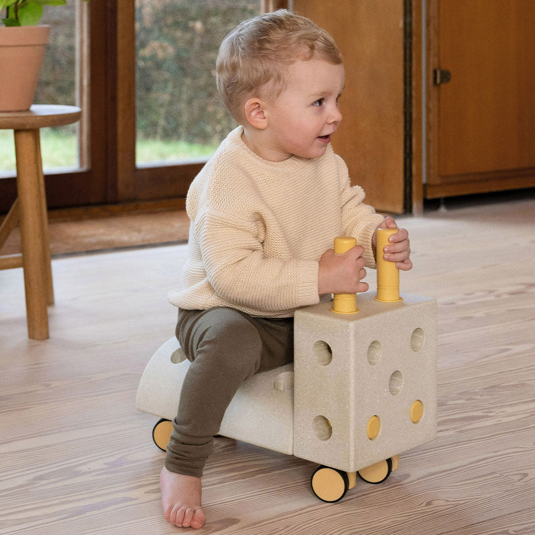 Een peuter zit op de Modu Tiny Ride loopwagen en kijkt naar de zijkant.