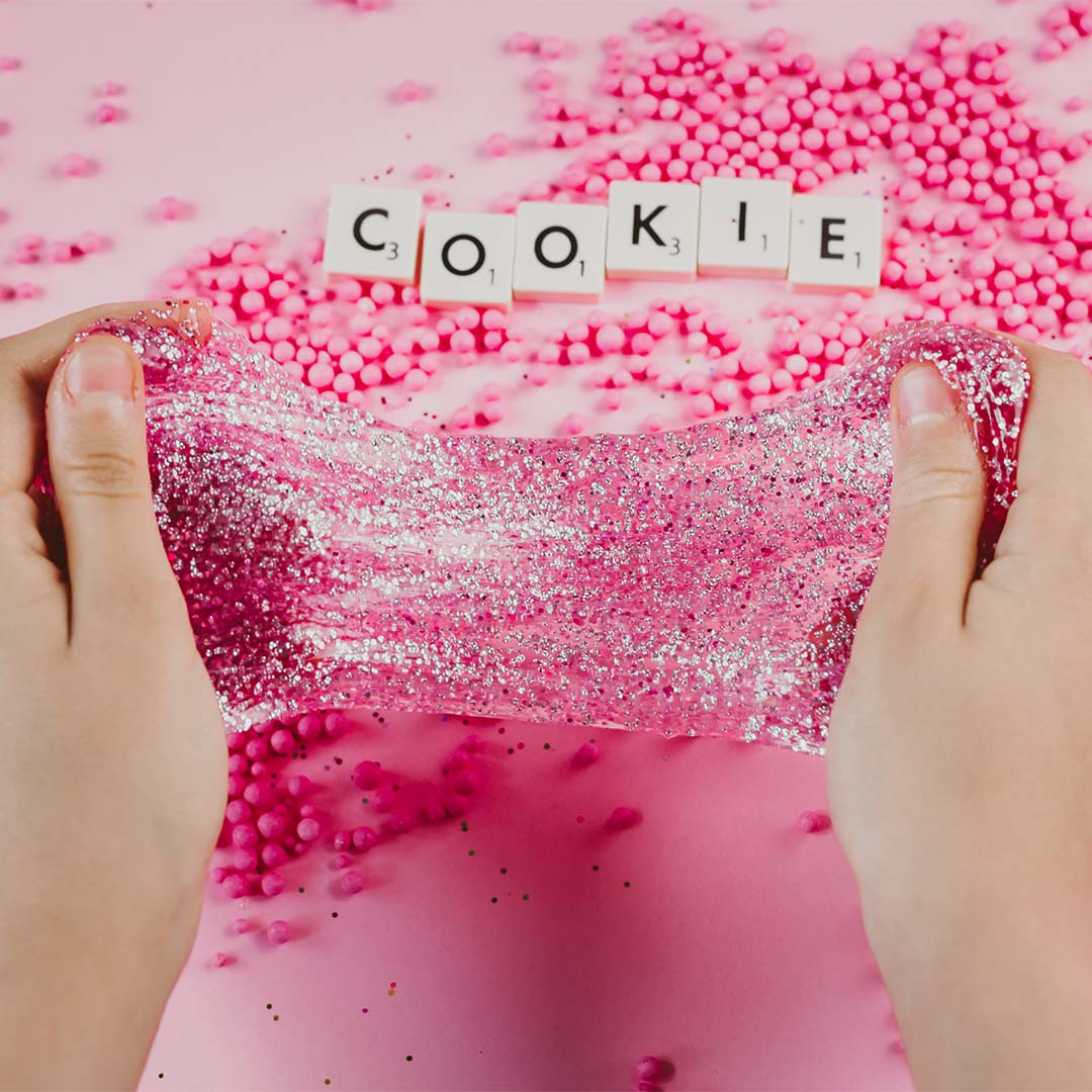 Handen rekken roze glitter slijm uit boven roze schuimballetjes en Scrabble-letters die 'COOKIE' spellen.