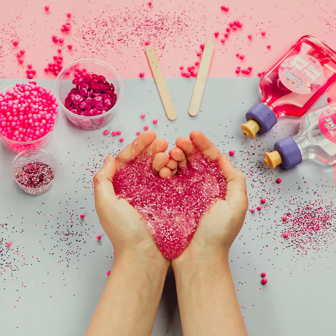 Handen vormen een hartje van roze Tuban glitter slijm op een roze-grijze ondergrond.