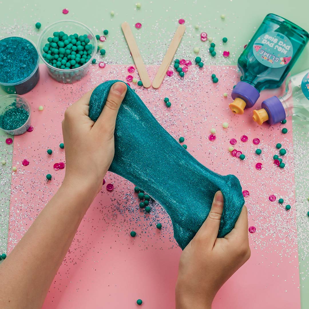 Twee handen spelen met uitgerekt blauw glitterslijm op een roze ondergrond.