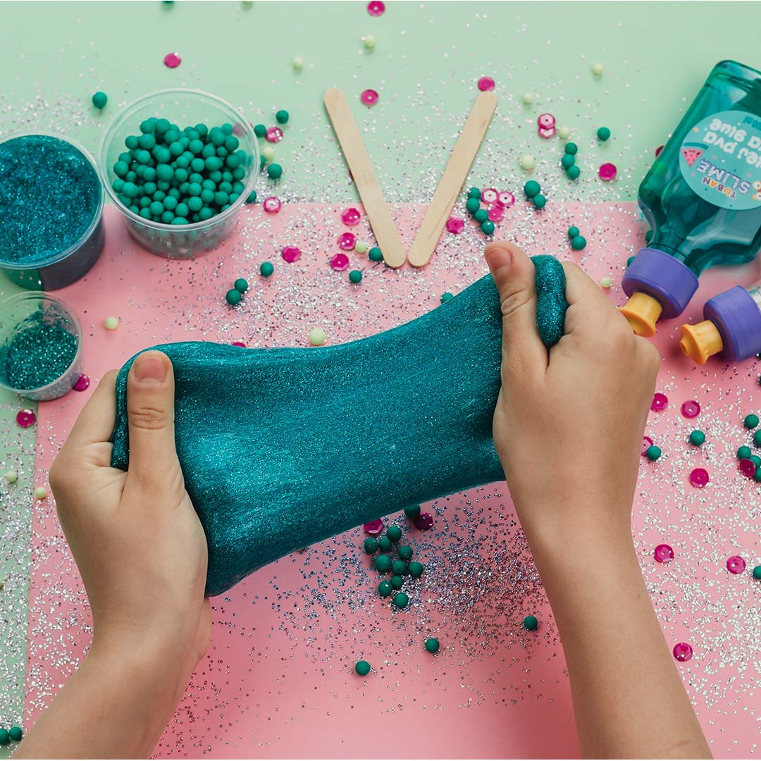 Twee handen rekken blauwe glitter slijm, met Tuban slijmbenodigdheden op de achtergrond.