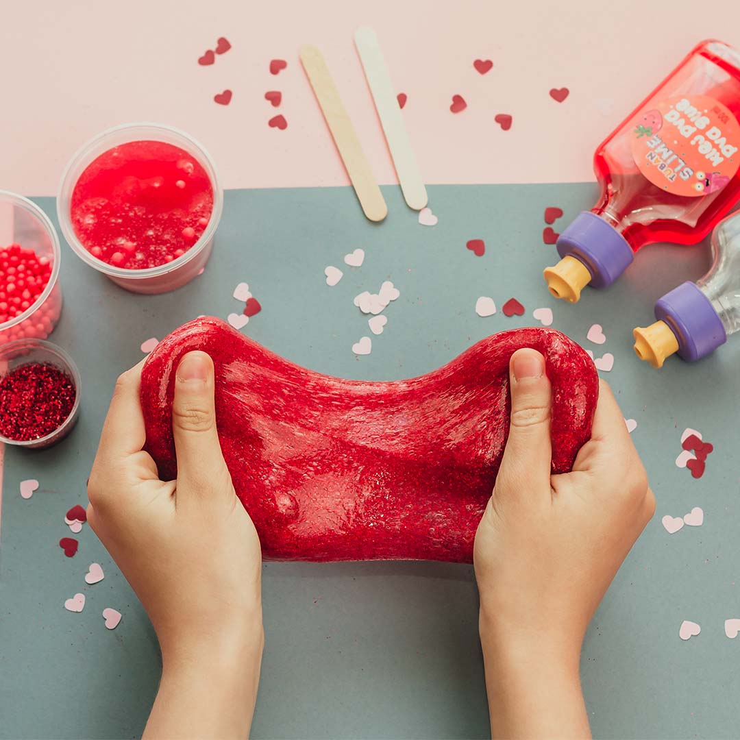 Kinderhanden rekken rood glitter slijm uit, met slijmbenodigdheden en hartjesconfetti op de achtergrond.