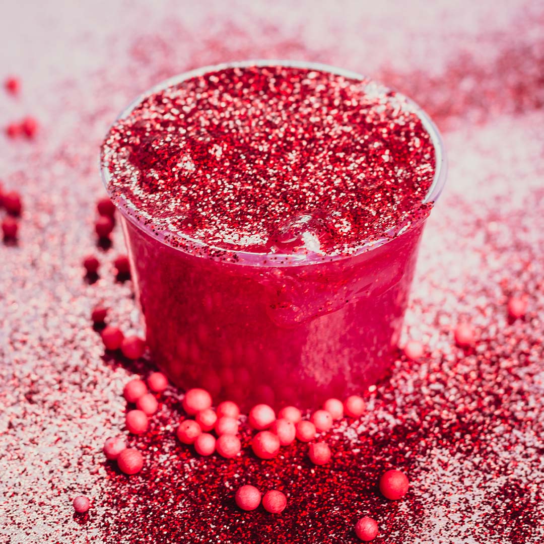 Detailfoto van een potje rood glitter slijm met losse glitters en rode balletjes eromheen.