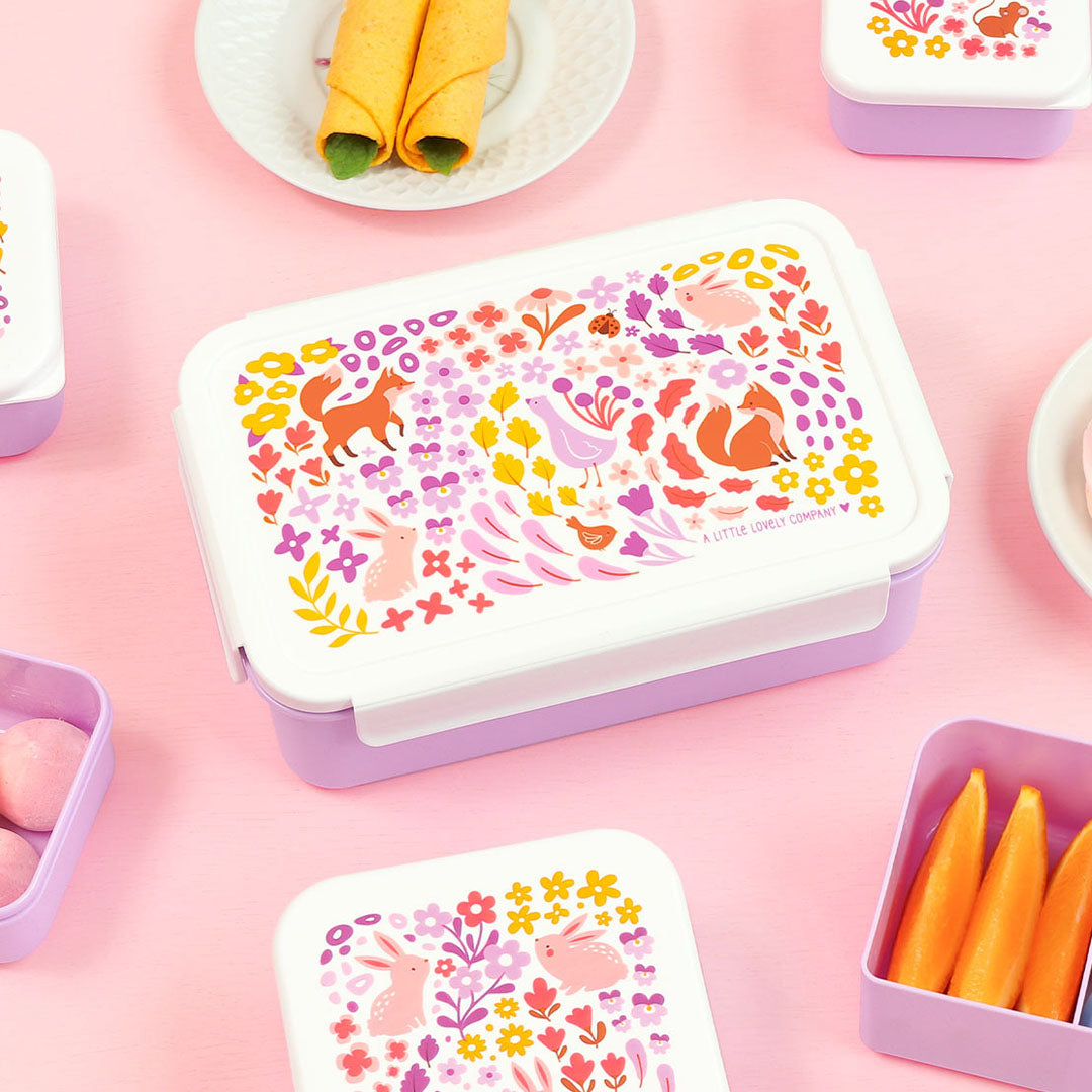 Een paarse bento broodtrommel met bloemen- en dierenpatroon op een roze ondergrond, omringd door kleinere containers en snacks.