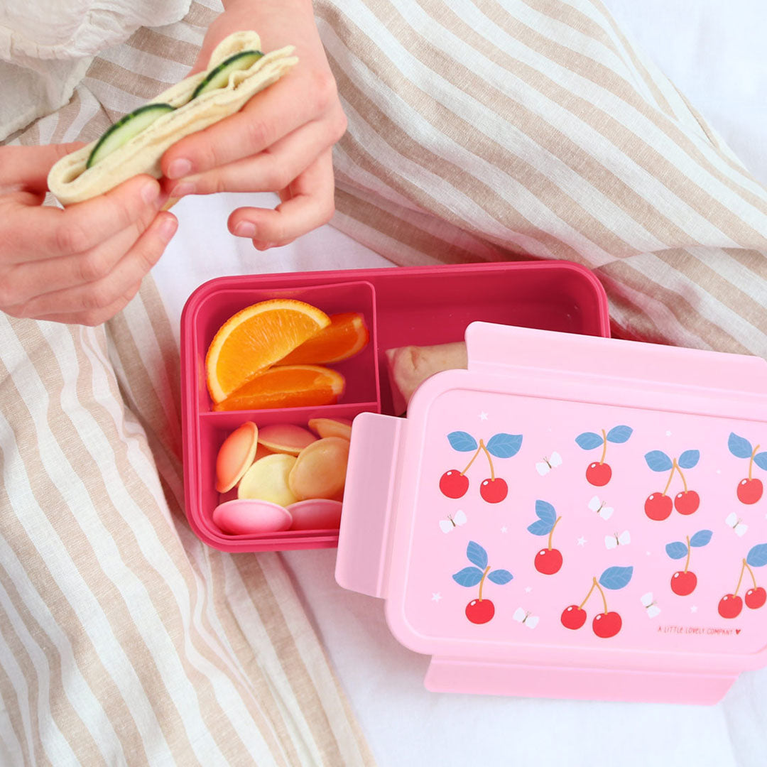 Een open roze a little lovely company bento broodtrommel met kersenpatroon, gevuld met fruit en snacks, naast handen die een wrap vasthouden.