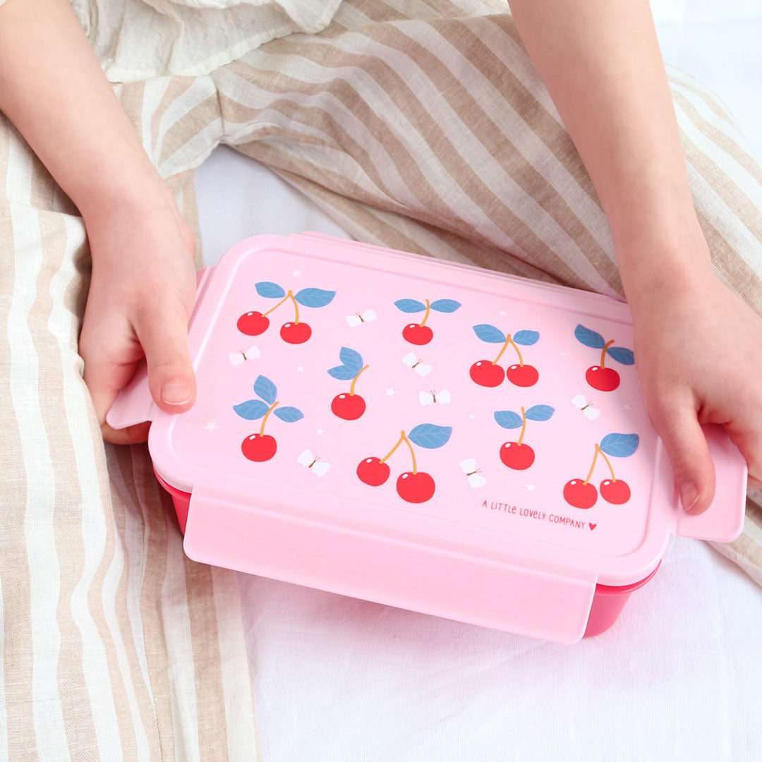 Kinderhanden houden een gesloten roze a little lovely company bento broodtrommel met kersenpatroon vast.