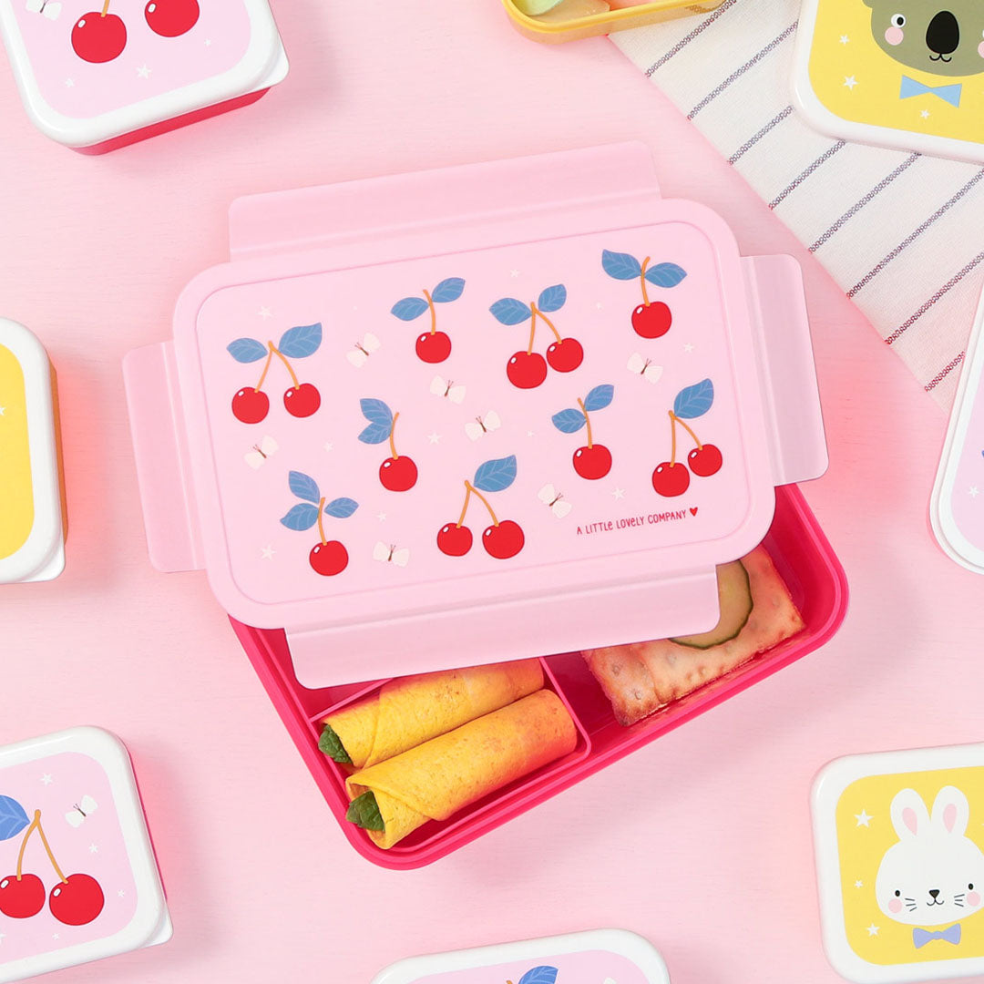 Een open roze a little lovely company bento broodtrommel met kersenpatroon, gevuld met eten, omringd door andere lunchboxen.