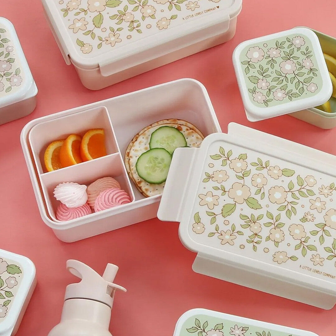A Little Lovely Company roze broodtrommel voor kinderen met bloesempatroon, gevuld met snacks en omringd door andere lunchboxen op een roze ondergrond.