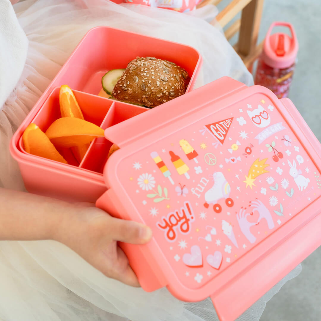 Kinderhanden openen de a little lovely company roze broodtrommel met 'fun' thema, gevuld met een broodje en sinaasappel.