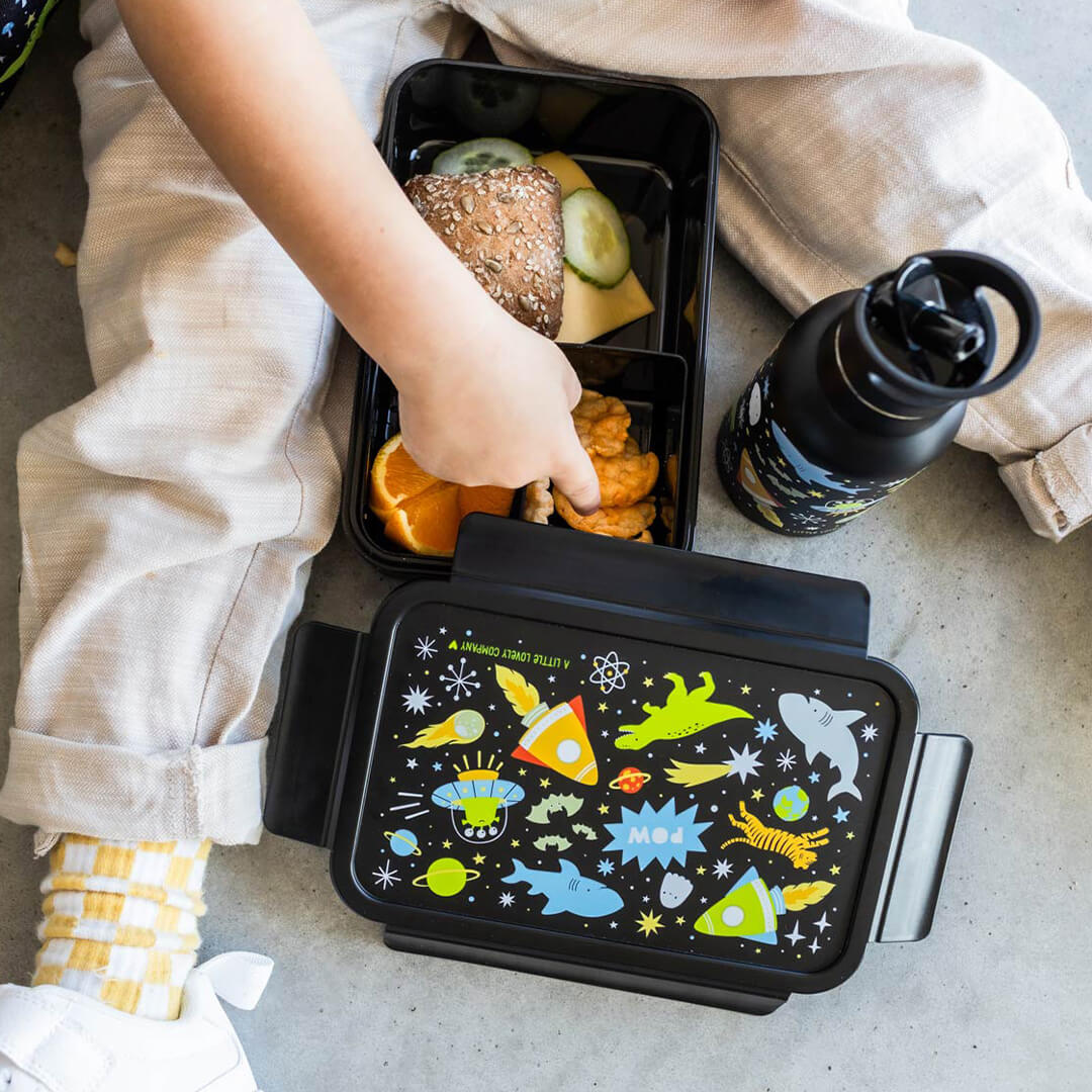 Een kinderhand pakt eten uit een open a little lovely company kinderbroodtrommel met galaxy-thema, naast een bijpassende drinkfles.