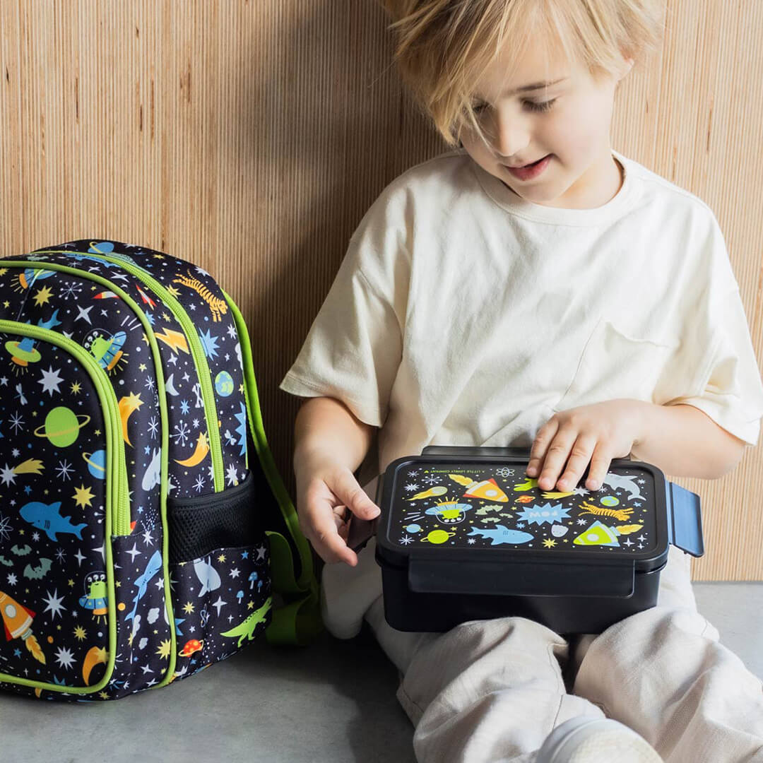 Een kind zit op de grond en houdt een a little lovely company kinderbroodtrommel met galaxy-thema vast, naast een bijpassende rugzak.