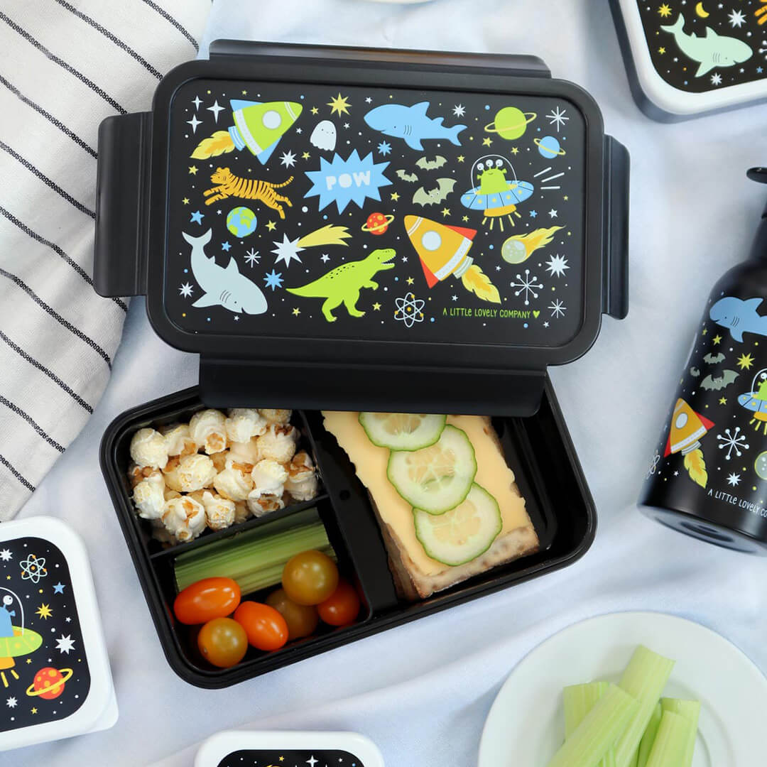 Een open a little lovely company kinderbroodtrommel met galaxy-thema, gevuld met popcorn, tomaatjes, selderij en een boterham met kaas en komkommer.